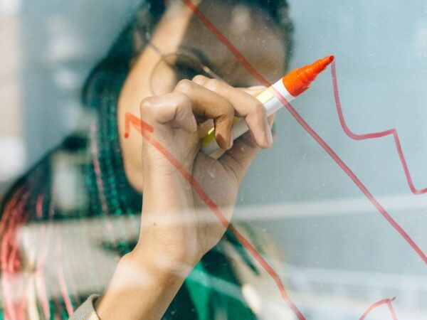 woman in a beige coat writing on a glass panel using a whiteboard marker