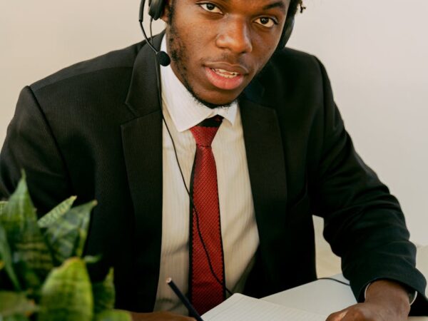 man in black suit writing on a notebook while seriously looking at the camera